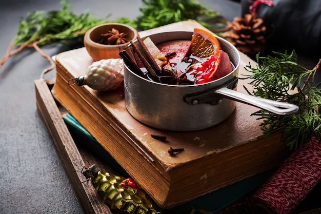 Mulled wine hot drink with citrus, apple, pomegranate and spices in aluminum casserole with vintage hristmas tree toys and Fir branch on concrete surface. Selective focuse.