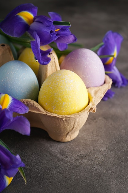 Multi colored Easter eggs in a cardboard box and fresh flower on a wooden surface, vertically. Close-up  Happy Easter concept.