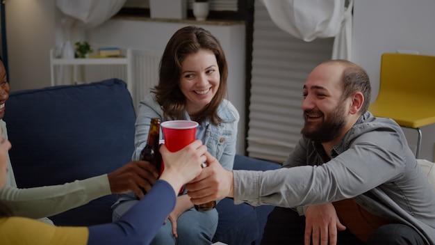 Multi cultural friends cheering beer bottles during wekeend party laughing while relaxing on sofa late at night. Group of mixed race people hanging out enjoying time spending together