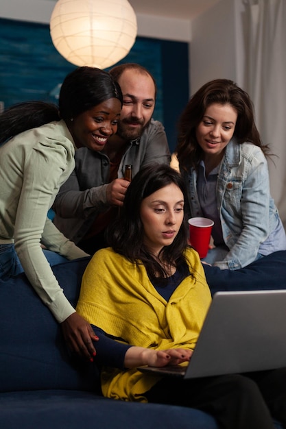 Multi-ethnic friends smiling while watching online comedy series on laptop computer during entertainment movie home night. Group of people enjoying hanging out together. Friendship concept