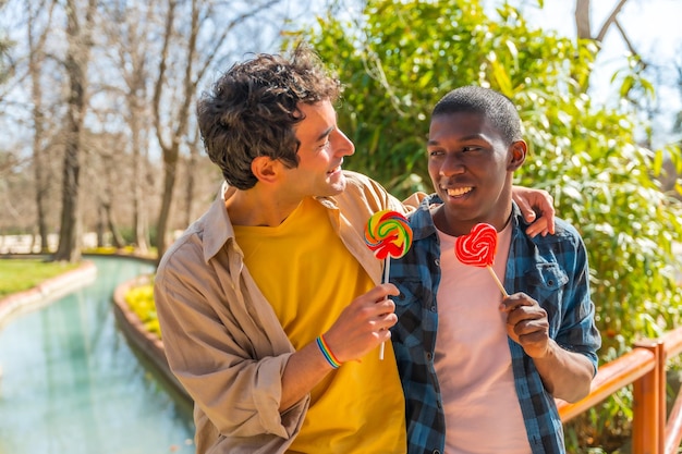 Multi ethnic gay male couple eating a lollipop lgbt concept having fun and smiling in a park