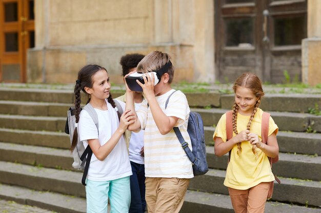 Multi-ethnic group of cheerful school children playing with VR glasses outdoors, focus on boy wearing gear, copy space