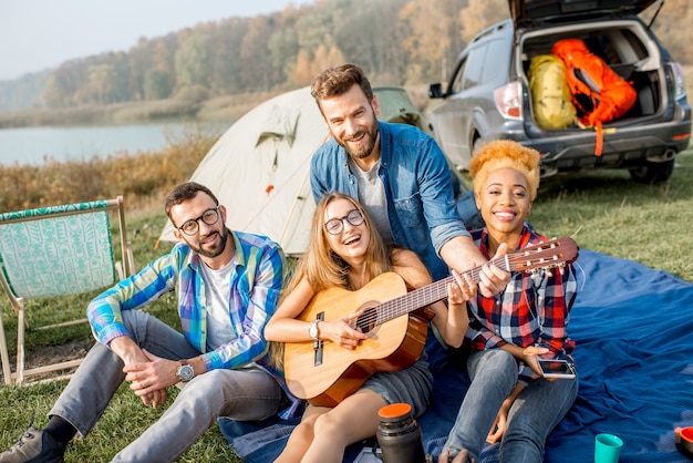 Multi ethnic group of friends dressed casually having fun playing guitar during the outdoor recreation with tent, car and hiking equipment near the lake