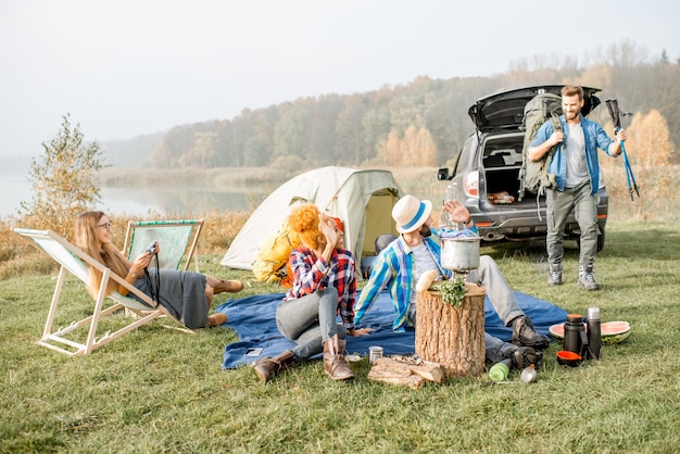 Multi ethnic group of friends dressed casually having a picnic during the outdoor recreation with tent, car and hiking equipment near the lake