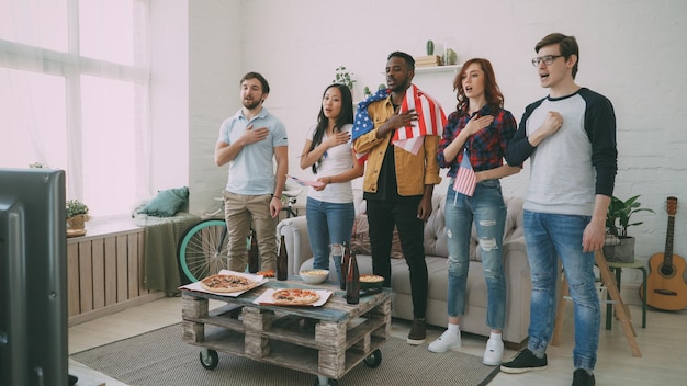 Multi ethnic group of friends sport fans singing national USA anthem before watching sports championship on TV together at home