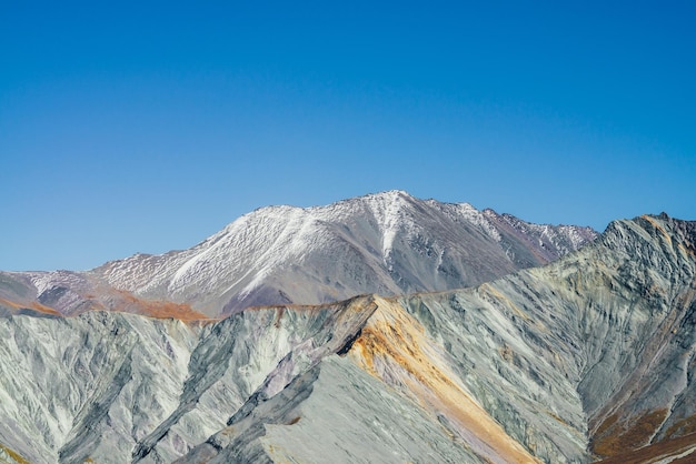 Multicolor autumn landscape with snow-covered mountain peak and gray rockies with orange and lilac tint. Spectacular colorful view to sharp mountain ridge. Motley mountain scenery in autumn colors.