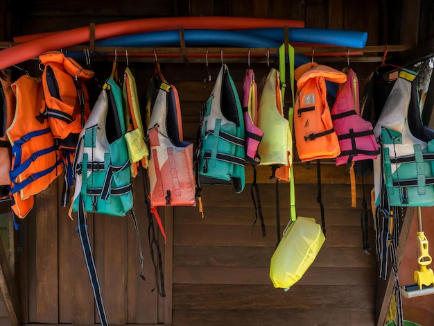 Multicolored life jacket vests are hung on rail on wood wall for resort guests who want to do water activities kayaking standup paddleboarding surfing and swimming summer holiday travel concept