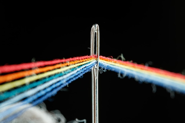 Multicolored threads for sewing in the form of a rainbow pass through an antique needle on a black background