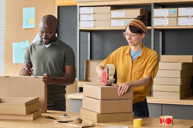 Multiethnic couple of workers packing parcels in cardboard boxes together before shipping