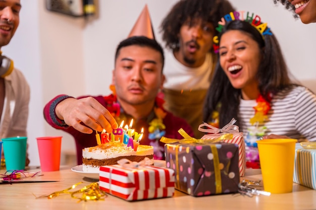 Multiethnic group of friends at a birthday party on the sofa at home with a cake and gifts Chinese man lighting the candles