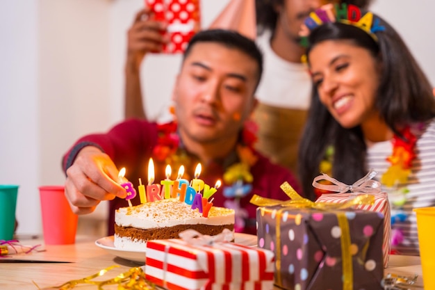 Multiethnic group of friends at a birthday party on the sofa at home with a cake and gifts lighting the candles