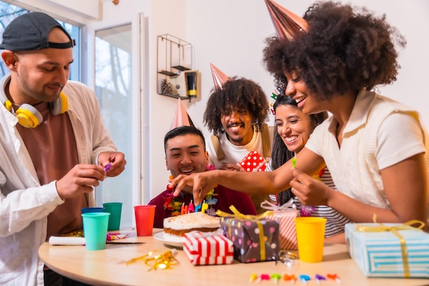 Multiethnic group of friends at a birthday party on the sofa at home with a cake and gifts placing the candles