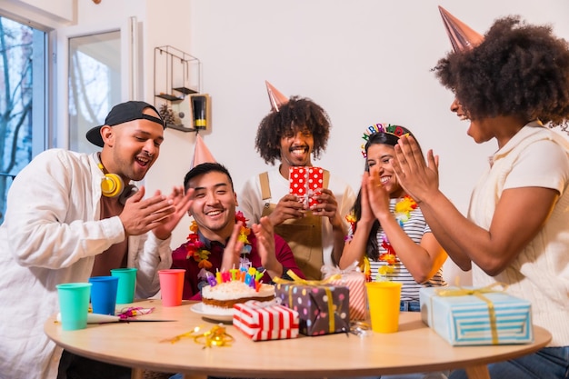 Multiethnic group of friends at a birthday party on the sofa at home with a cake and gifts smiling placing the happy brithday candles