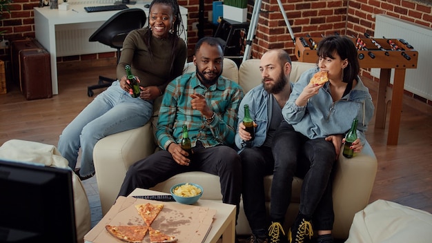 Multiethnic group of friends clinking beer bottles at house party, making toast and watching film on tv. Happy people enjoying movie on television channel with alcohol drinks.