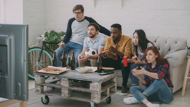 Multiethnic group of friends sports fans watching football championship on TV together eating pizza and drinking beer at home