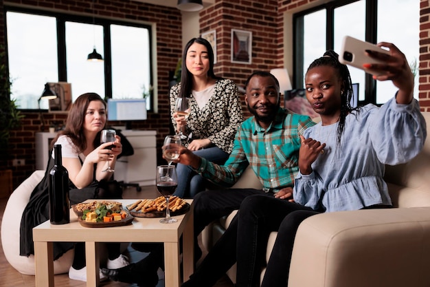 Multiethnic group of friends taking selfie photo together while at wine party in living room. Happy smiling adults at home enjoying time together while drinking alcoholic beverages and taking pictures