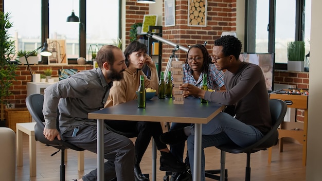 Multiethnic group of people having fun with tower pieces, playing board games with square building and wooden blocks on table. Friends laughing and enjoying leisure pasttime activity.