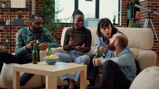 Multiethnic group of people talking about friendship and drinking beer from bottles to have fun at home gathering. Friends laughing and enjoying weekend activity together, relaxing.