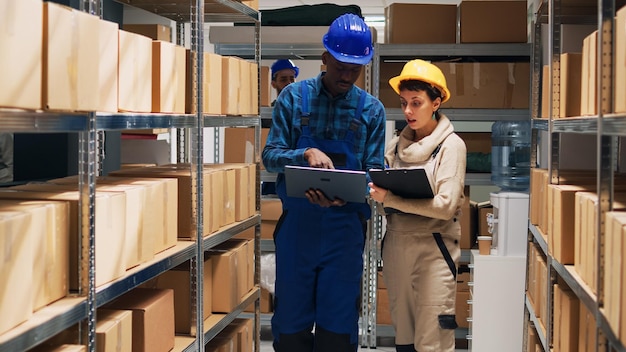 Multiethnic team of employees working with goods and checking stock inventory on laptop, cargo in storage room. Depot workers analyzing industrial goods to plan distribution and shipment.