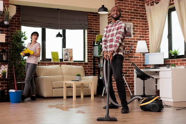 Multiracial couple happily cleaning at home, african american man vacuuming the whole place while woman dusts the shelves. Working as a team to keep house clean and tidy.