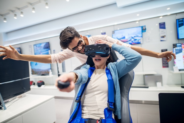 Multiracial couple having fun with VR goggles while girl sitting in the chair in the tech store. Customer's service. Shopping time.