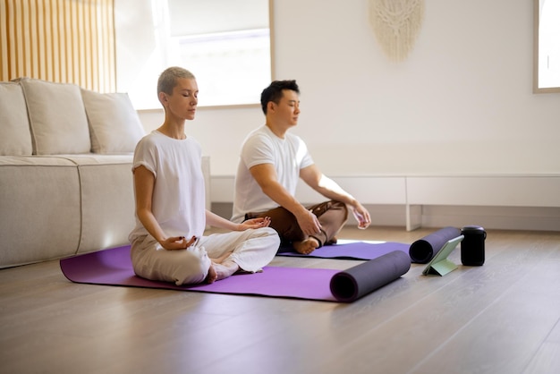 Multiracial couple practising yoga and meditating while sitting on fitness mats at home. Concept of healthy lifestyle. Idea of domestic hobby and leisure. Asian man and caucasian girl with closed eyes