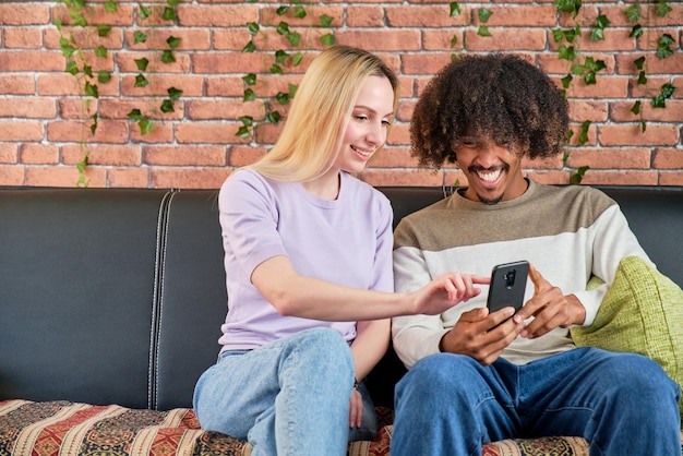 Multiracial couple sitting on the sofa at home looking at go on their smart phone smiling