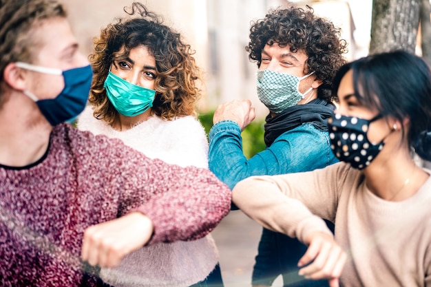 Multiracial friends greeting with elbow bump wearing face protection - Focus on left woman