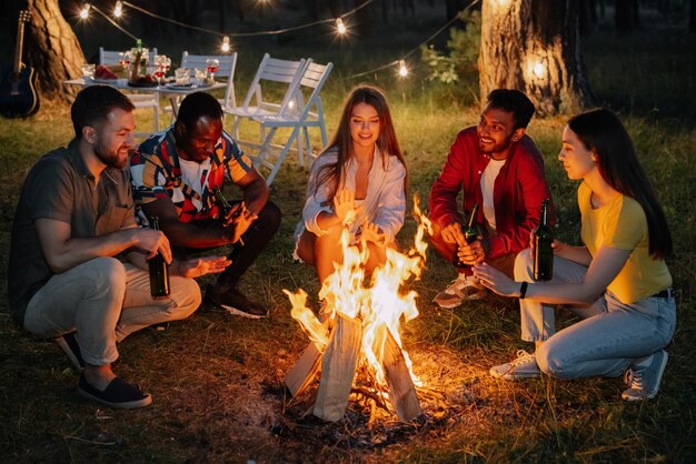 Multiracial friends sitting around the fire drinking beer and having fun at the party