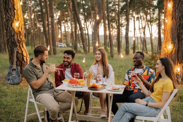 Multiracial group of happy friends sitting around table drinking wine talking at dinner party