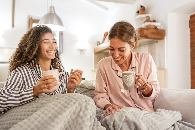 multiracial homosexual female couple having fun at home sitting at the sofa drinking a cup of tea under a blanket - Two beautiful young women bonding each other