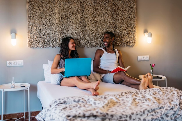 A multiracial married couple smiles in bed while she shows him something on the laptop