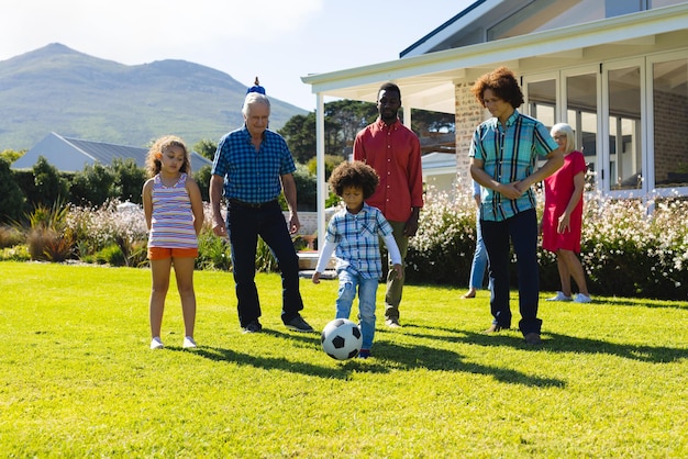 Multiracial multigeneration family looking at boy playing soccer on grassy field in yard in summer