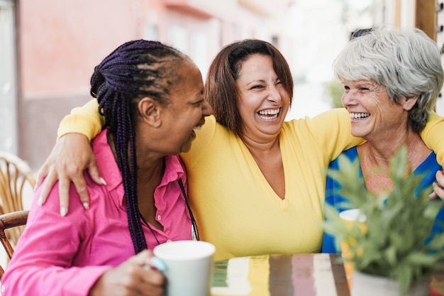 Multiracial senior friends meet and chat at bar outdoor while drinking coffee together at bar restaurant - Focus on center woman face