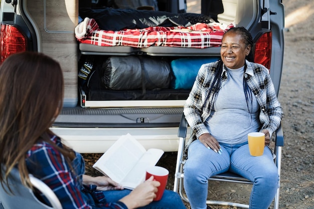 Multiracial women friends having fun camping with camper van while reading and drink coffee outdoor  Focus on african senior face