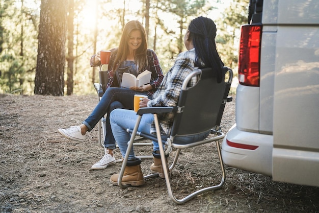 Multiracial women friends having fun camping with camper van while reading and drink coffee outdoor  Focus on left senior hand holding book