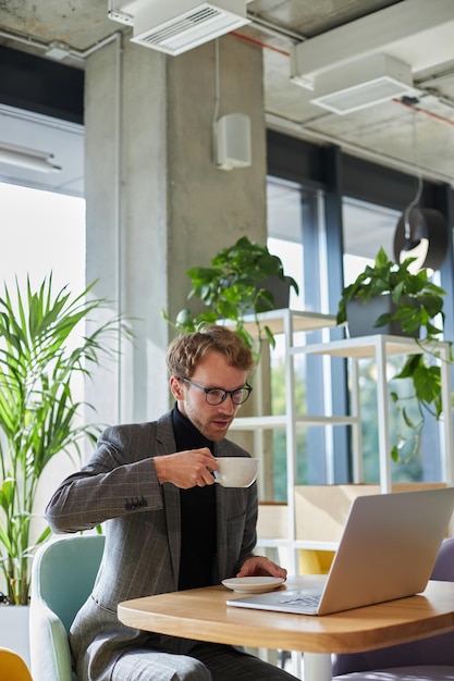 Multitasking confident businessman drinking coffee while telecommuting on laptop from modern office