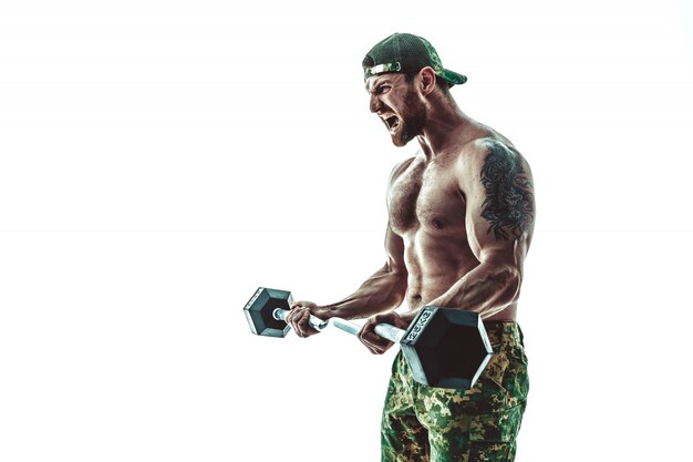 Muscular athlete bodybuilder man in camouflage pants with a naked torso workout with dumbbell on white