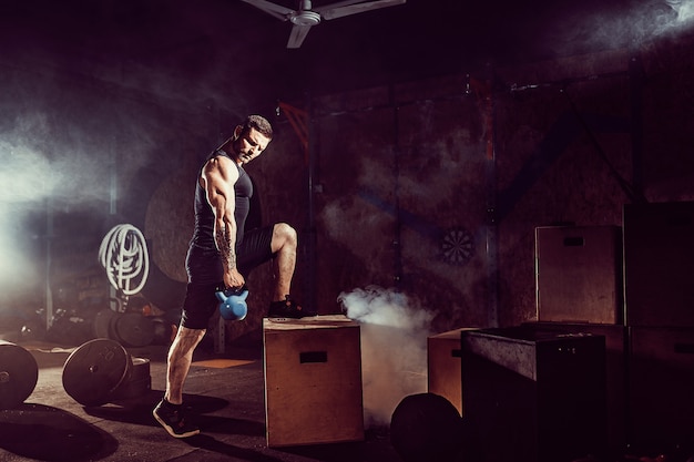 Muscular attractive caucasian bearded man lifting two kettlebells in a gym. Weight plates, dumbbell and tires in background.