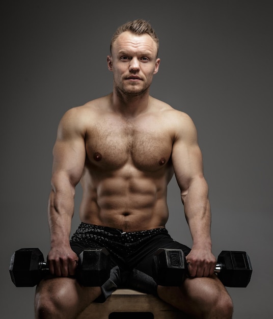 Muscular bodybuilder guy sitting on chair and doing exercises with dumbell on grey background