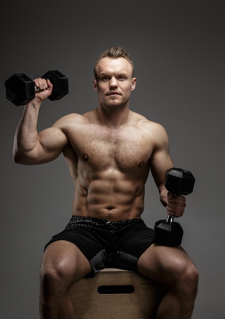 Muscular bodybuilder guy sitting on chair and doing exercises with dumbell on grey background