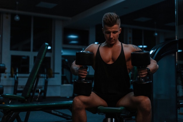 Muscular bodybuilder man sits with dumbbells and trains in the gym. Sporty lifestyle