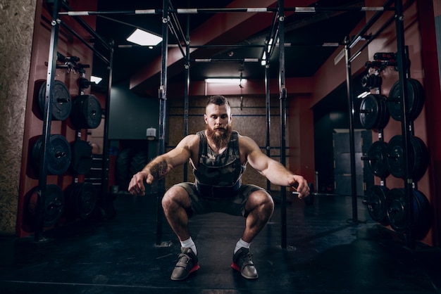 Muscular caucasian bearded man doing squats in military style weighted vest in crossfit gym.