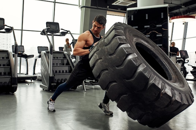 Muscular fitness man moving large tire in gym center concept lifting workout training