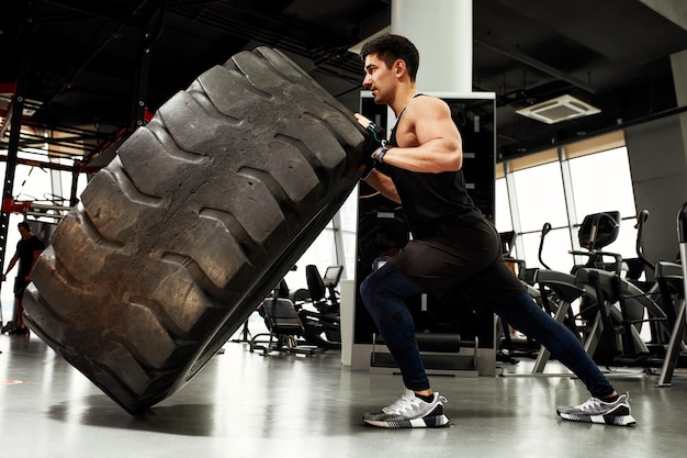 Muscular fitness man moving large tire in gym center concept lifting workout training