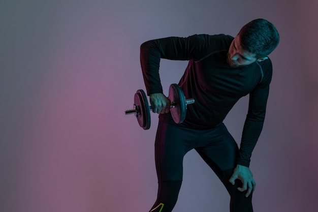 Muscular guy in sportswear working out with dumbbell, pumping up shoulder muscles with heavy weight on grey studio wall. Sport, bodybuilding and active lifestyle concept