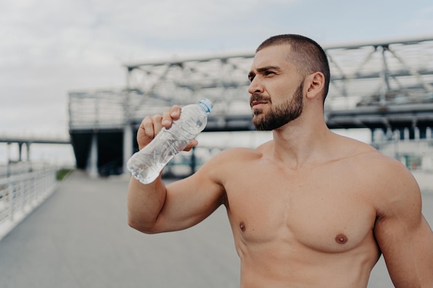 Muscular guy takes a break drinks water cares about hydration poses outside after jogging