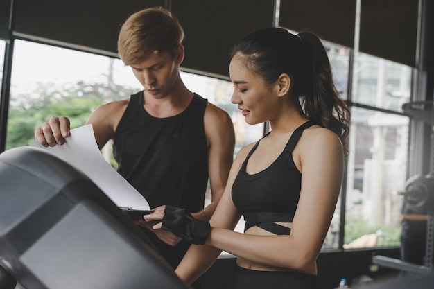 Muscular handsome caucasian personal trainer man discussion with young asian woman slim body exercising his legs cardio running on treadmill in fitness gym for good healthy