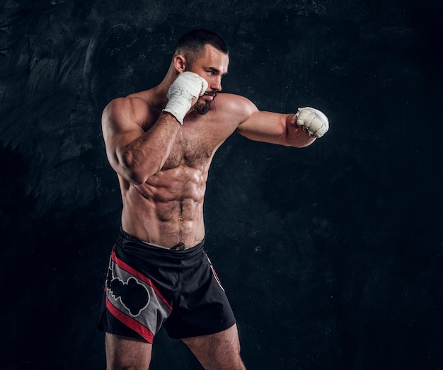 Muscular handsome fighter with naked torso is demonstraiting his power at dark photo studio.