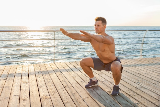 Muscular handsome man with naked torso doing
pre-workout squat exercises on the beach at sunrise